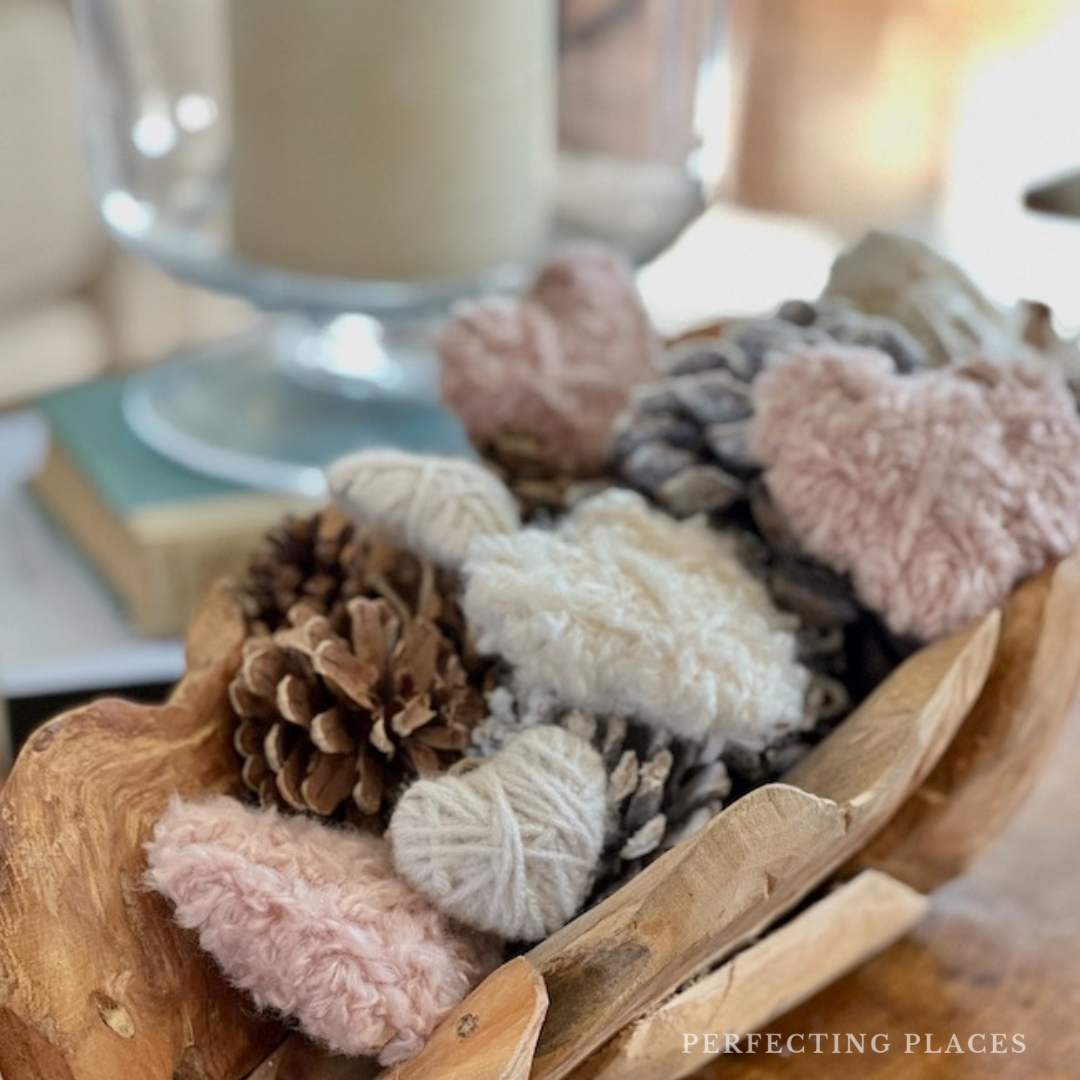 A wooden bowl on the table holds a mix of pinecones and heart-shaped woolen decorations in soft pink and cream, perfect for adding cozy home touches. In the background, there's a glass candle holder accompanied by a stack of favorite books.