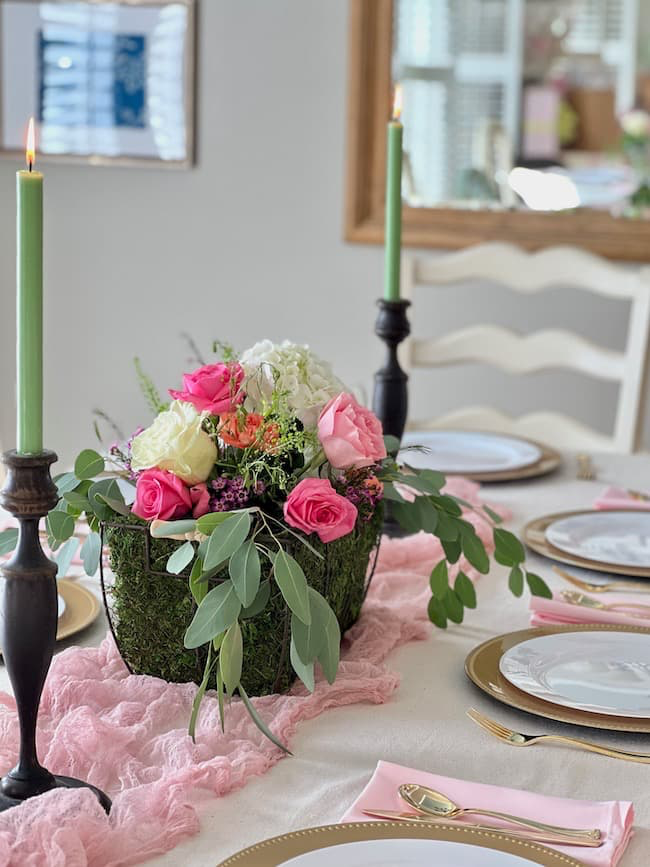 A dining table decorated with a light pink table runner, a floral centerpiece with pink and white roses, and greenery. There are two lit green candles on black holders and white plates set on gold-rimmed chargers with pink napkins.