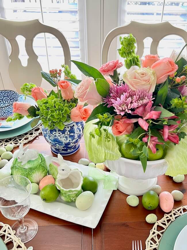 A dining table decorated for spring with two floral arrangements featuring pink roses and green hydrangeas in patterned vases. A ceramic bunny sits among pastel green and white eggs. The background includes white chairs and a window.