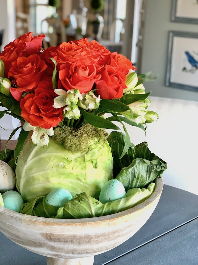 A decorative centerpiece features vibrant orange roses and white flowers arranged in a green cabbage. Surrounding the cabbage are light blue speckled eggs nestled in cabbage leaves, all placed in a rustic wooden bowl on a gray table.