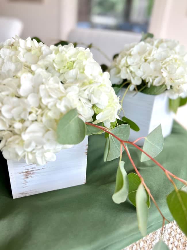 White hydrangea flowers in rustic white wooden boxes sitting on a green fabric surface. Eucalyptus branches are draped elegantly across the arrangement, with a blurred background.