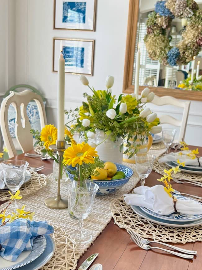 A dining table set for a meal with white plates, blue napkins, and glasses. A floral centerpiece featuring white tulips and yellow flowers complements additional small bouquets. A wicker placemat and a decorative mirror with a wreath are in the background.