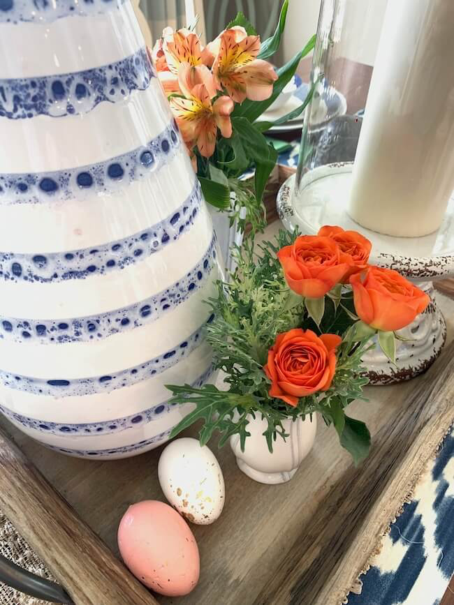 Ceramic vases with blue and white stripes, orange lilies and roses, and two decorative eggs, one pink and one speckled, arranged on a wooden tray.