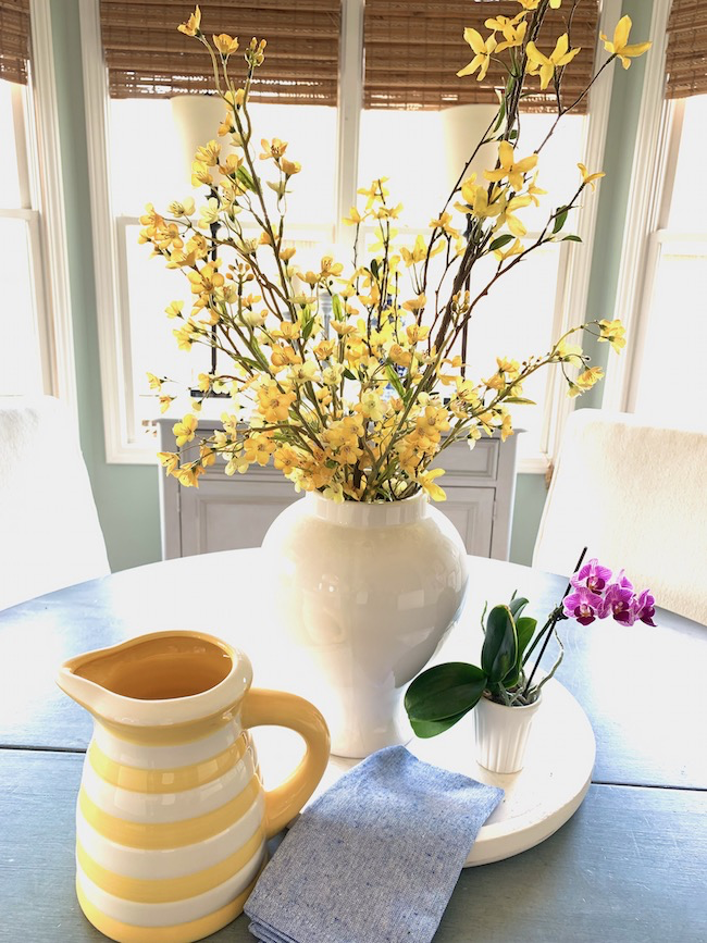 A white vase with yellow forsythia blossoms sits on a round table. A small potted orchid and a yellow-striped pitcher are nearby on a white tray, along with a folded blue cloth. Sunlight streams in through the windows in the background.