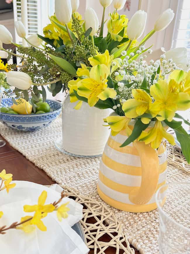 A table decorated with two vases of yellow and white flowers, including tulips and alstroemeria. The vases rest on a woven table runner. A blue and white bowl filled with limes is visible in the background.