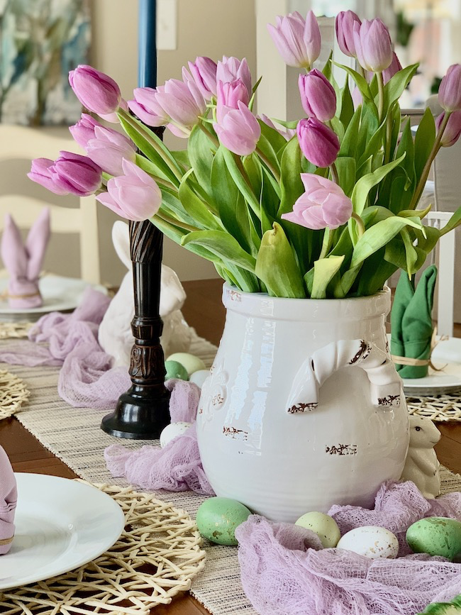 A table is decorated with a white vase filled with pink and purple tulips. There are pastel-colored eggs, plates, and napkins shaped like rabbits on a woven placemat. Two candles stand in the background. The setting is bright and festive.