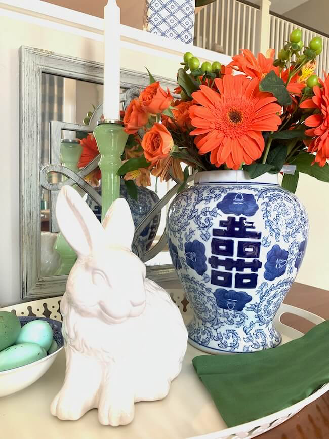 A white ceramic rabbit sits on a tray next to a blue and white vase filled with orange flowers and greenery. Behind it, a green candle in a holder is visible. The setting is indoors with a mirror reflecting part of the arrangement.