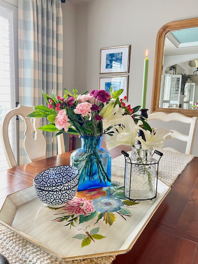 A dining table with a floral centerpiece in a blue vase, a lit green candle, and a decorative bowl on a flower-patterned tray. A large mirror, framed photos, and curtains are in the background.