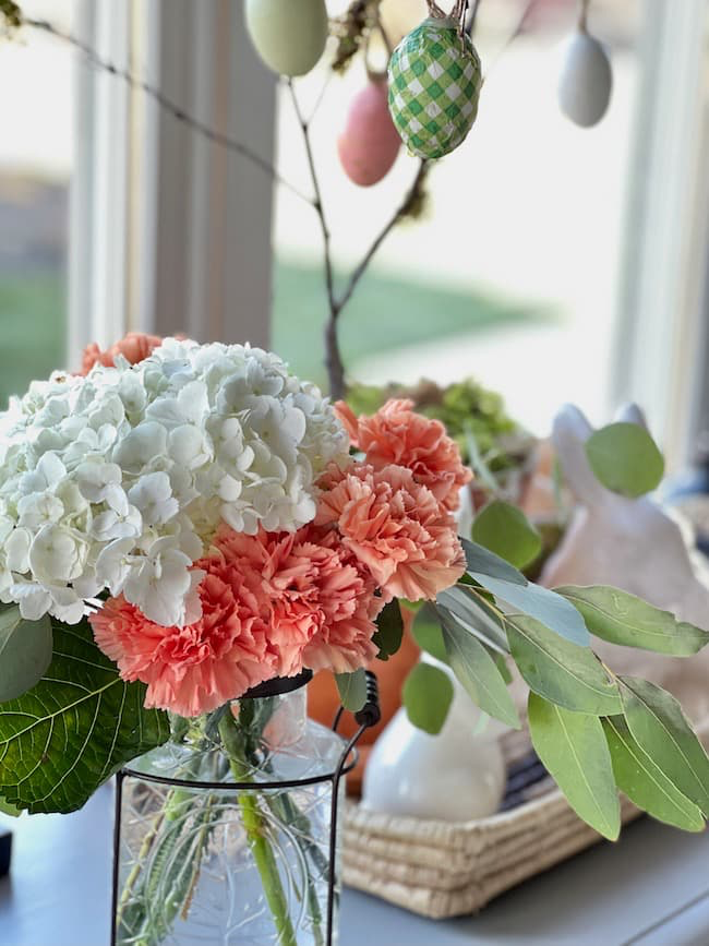 A bouquet of white hydrangeas and peach carnations in a glass vase sits on a table. Green leaves surround the flowers. Decorative eggs hang in the background with a ceramic rabbit and wicker basket nearby. A window offers a bright view outside.