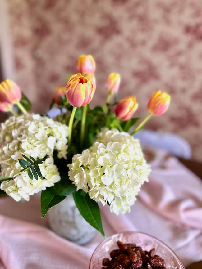 A vase holds a bouquet of pink and yellow tulips paired with white hydrangeas. The arrangement sits on a table covered with a light pink cloth. In front, there is a bowl of nuts. The background features a floral-patterned wall.