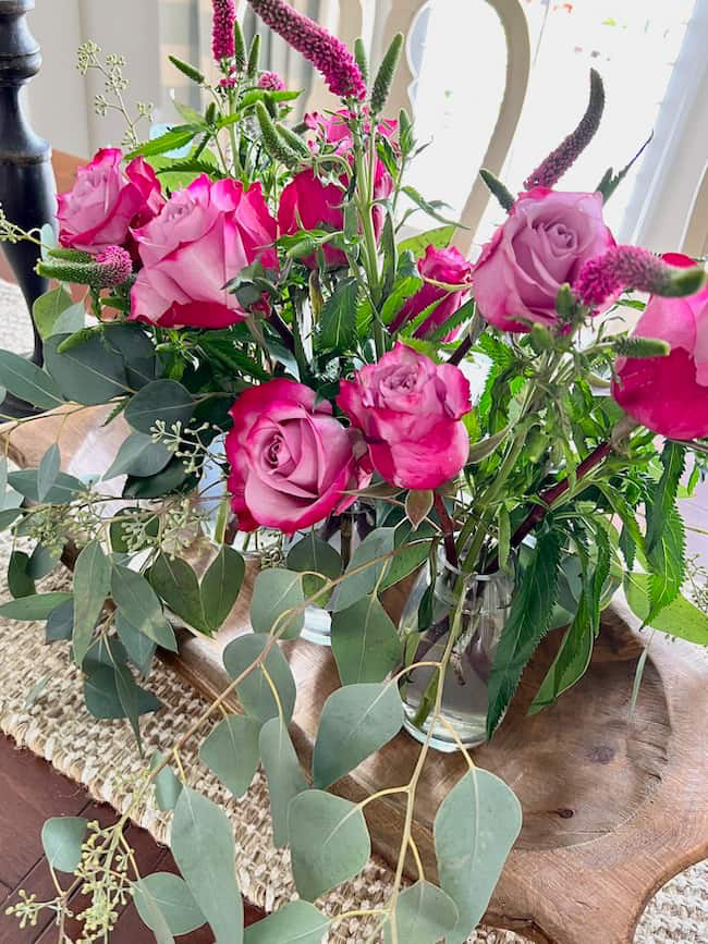 A bouquet of pink roses and greenery in clear vases on a wooden tray. The arrangement includes eucalyptus leaves and purple Veronica flowers, placed on a woven burlap table runner.