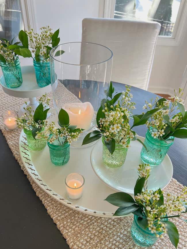 A dining table centerpiece features small green glass jars filled with white flowers and greenery. Candles, including a lit flame candle inside a glass hurricane, are arranged on a white tray atop a woven table runner.