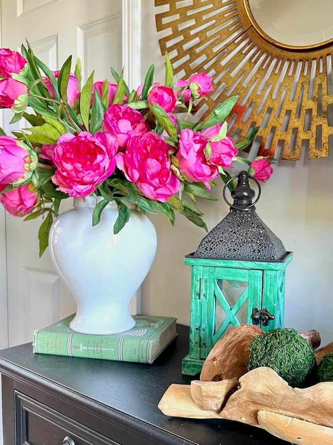 A white vase with vibrant pink flowers is placed on two green books. Next to it, a turquoise lantern and decorative wooden and green moss pieces rest on a dark table. A gold-framed mirror hangs in the background.