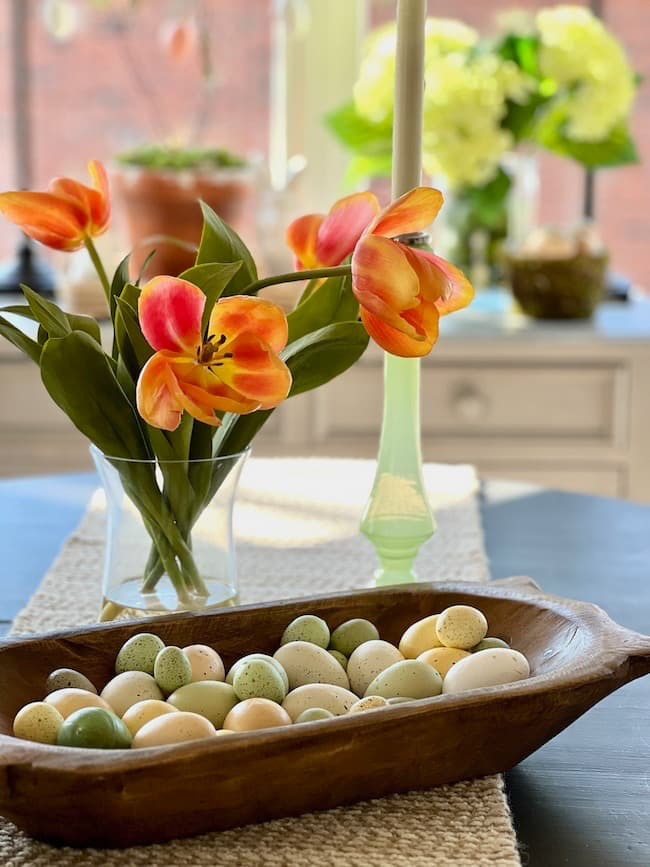 A wooden bowl filled with speckled eggs sits on a table runner. Beside it, a glass vase holds bright orange tulips, and a tall green candle holder stands nearby. In the background, potted plants are visible on a sunny window sill.