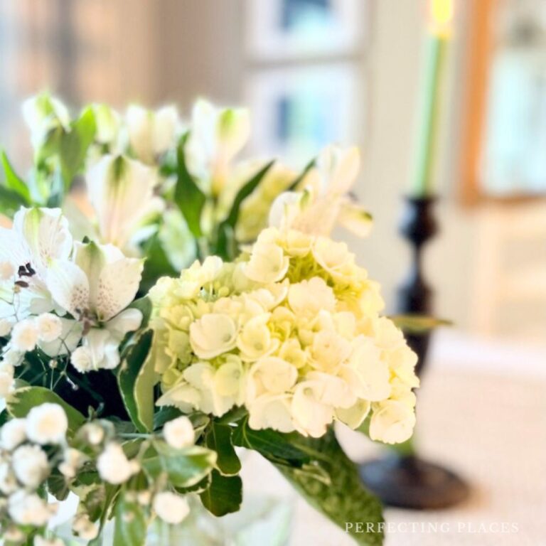 A close-up of a floral arrangement featuring white hydrangeas and other white flowers with green leaves in a vase. A lit green candle on a black candlestick holder is in the blurred background.