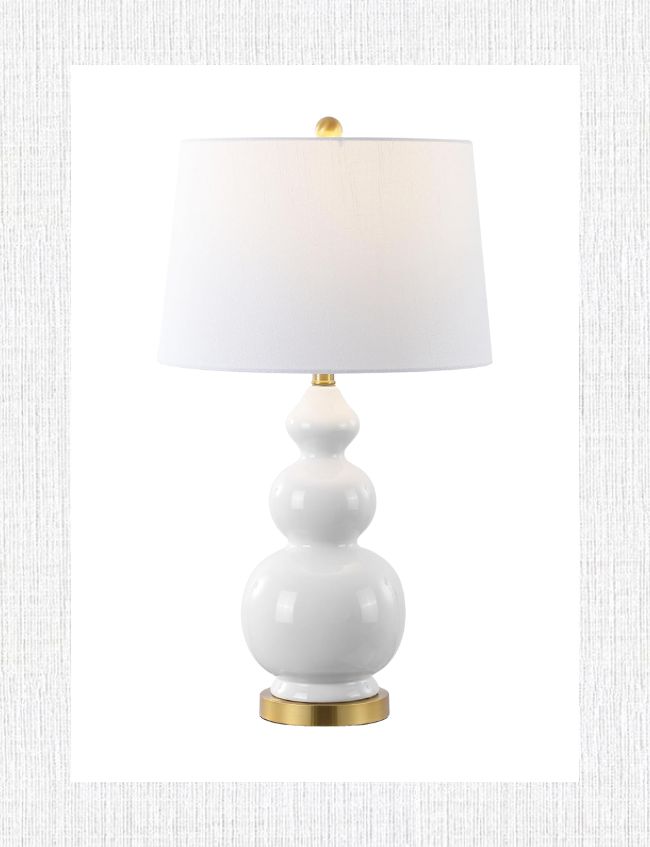 A white table lamp with a stacked, rounded ceramic base and a white cylindrical shade complements any spring decor. The base features three bulbous sections with gold accents, perfect for an entryway or foyer against a textured light gray background.