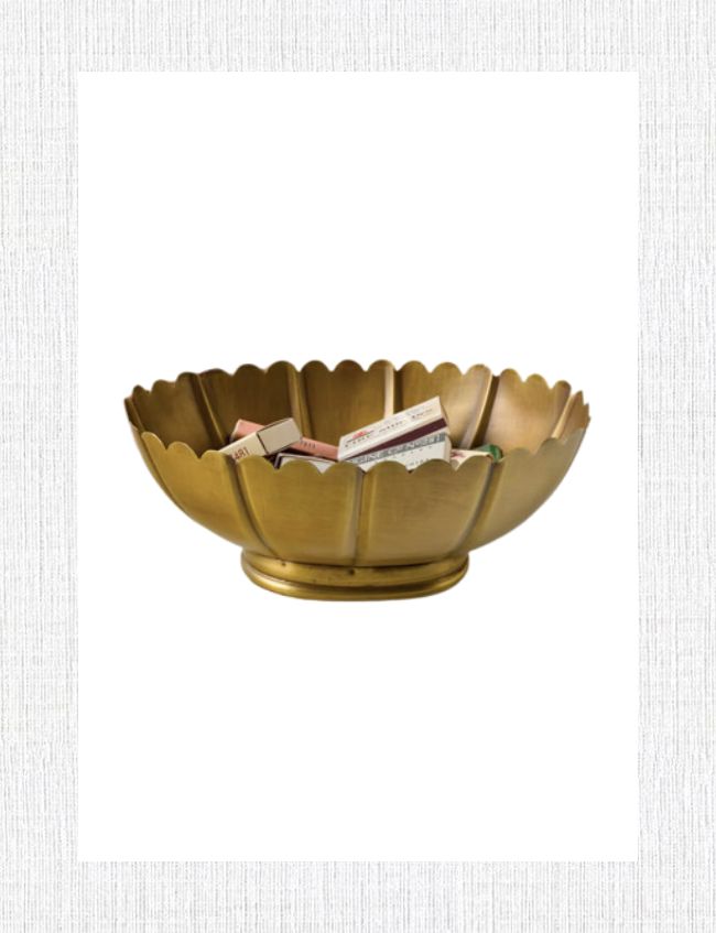 A gold-toned, scalloped metal bowl filled with an assortment of small matchboxes makes for perfect foyer styling. This elegant piece, with its pedestal base, stands gracefully against a plain white background, adding a touch of charm to your entryway decor.