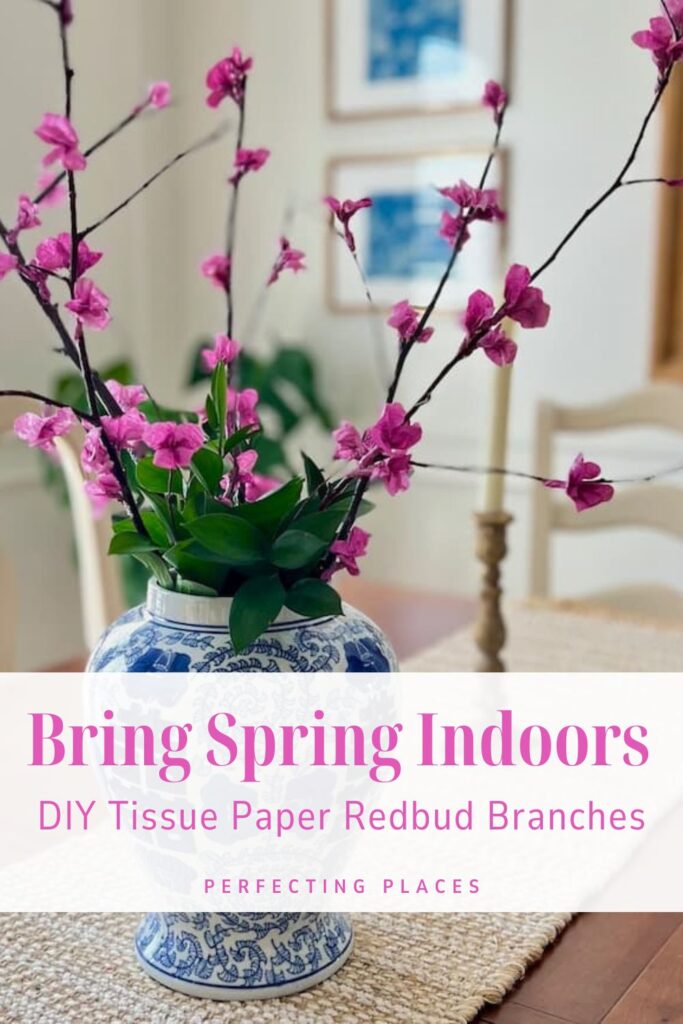 A blue and white vase with DIY tissue paper flowering branches and green leaves sits on a table. The overlay reads, "Bring Spring Indoors: DIY Tissue Paper Redbud Branches." In the background, a chair and framed pictures complete the charming scene.