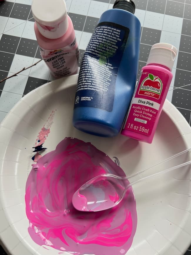 A paper plate with mixed pink and blue paint sits on a checkered surface. Nearby are three bottles: a small bottle of light pink paint, a medium bottle of blue paint, and a bright pink one. A plastic spoon rests on the plate, ready to create art inspired by tissue paper flowering branches.