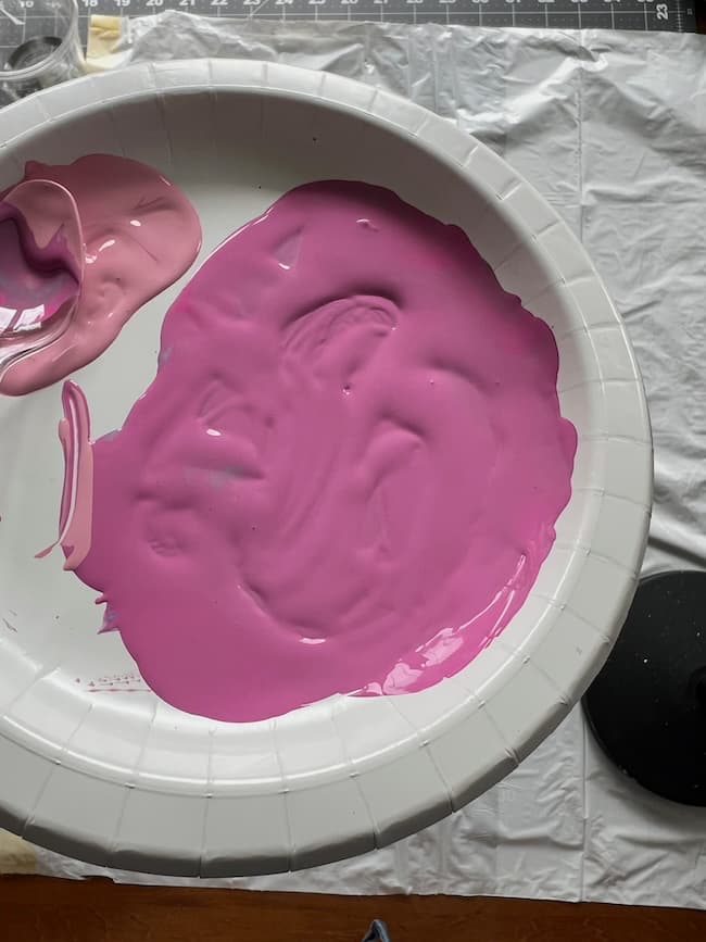 A white paper plate with vibrant pink paint smeared across it, featuring a lighter shade of pink in the corner, resembles delicate tissue paper. The background includes a black and white cutting mat and a black circular object, evoking the subtle elegance of flowering branches.