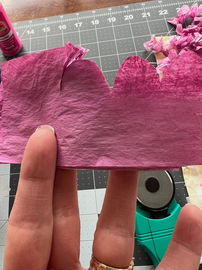 A hand clutches a piece of crinkled pink crepe paper cut into a wavy pattern, resembling delicate tissue paper. In the background, a cutting mat with a grid, along with tools and materials for crafting beautiful flowering branches, sets the creative scene.