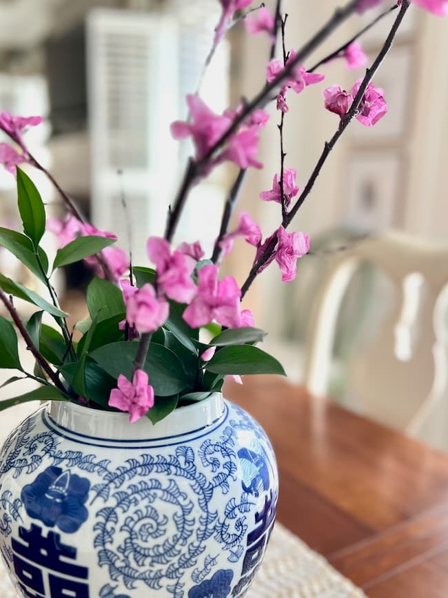 Blue and white ceramic vase with intricate dragon designs, holding tissue paper flowers alongside branches of vibrant pink blossoms and green leaves, placed on a wooden table with a softly blurred background.