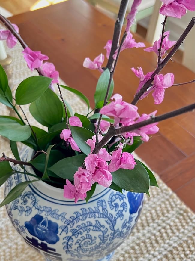 A decorative vase with intricate blue designs holds branches adorned with vibrant pink tissue paper flowers and green leaves, placed on a wooden surface with a woven mat beneath.