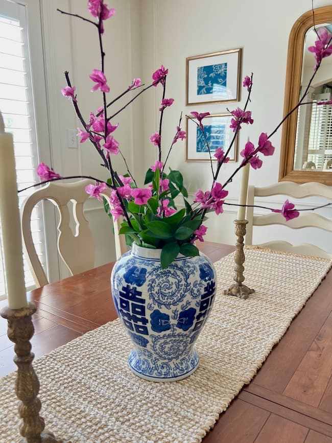 A blue and white vase with intricate patterns holds flowering branches crafted from tissue paper on a wooden dining table. The table features a woven runner and two candlesticks. In the background, a mirror and framed artwork adorn the cream-colored wall.