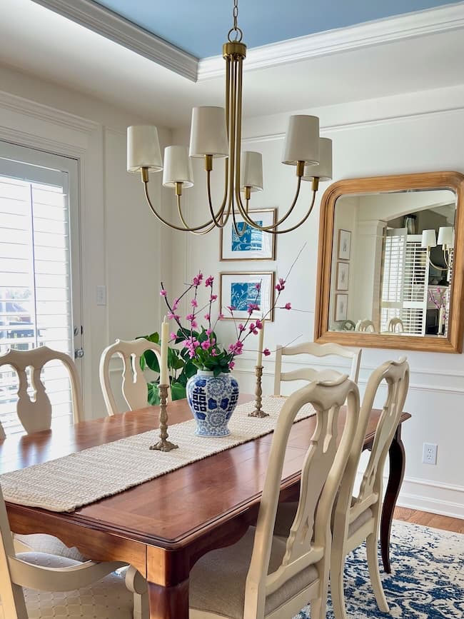 A dining room with a wooden table and cream-colored chairs. A blue and white vase with tissue paper flowers is on the table, alongside candlesticks. A large mirror and artwork adorn the white walls, while a chandelier hangs above. Flowering branches are delicately arranged on a blue rug below.