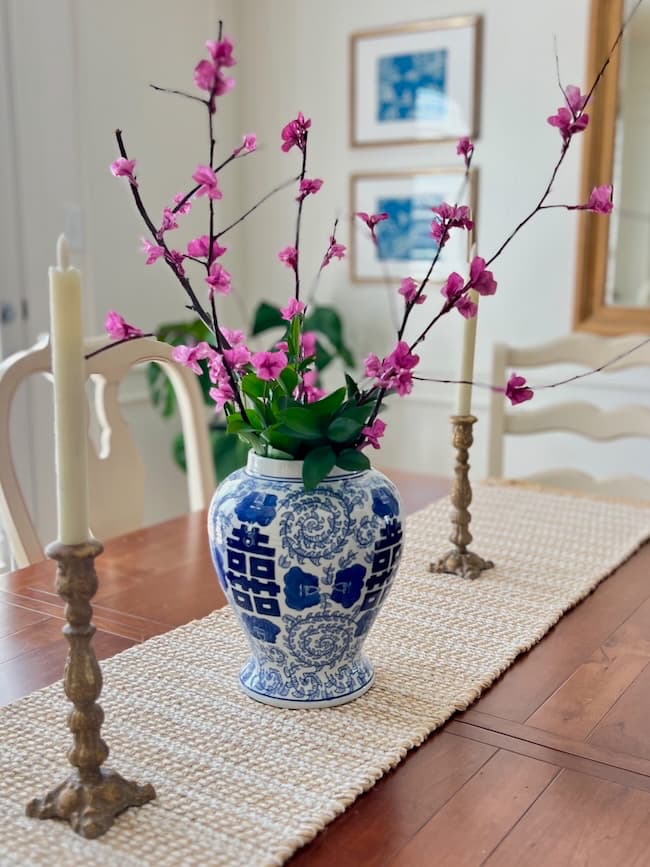 A blue and white vase with floral designs holds delicate branches of tissue paper pink blossoms. Positioned on a textured beige table runner on the wooden dining table, two tall white candles in brass holders flank the vase. Framed pictures adorn the wall in the background.