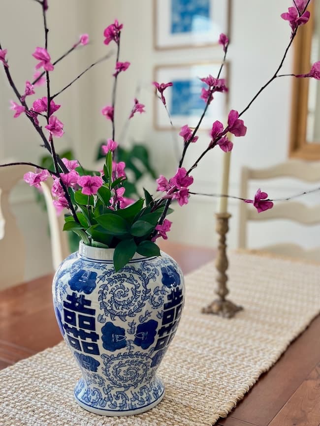 A blue and white floral vase holds branches adorned with bright pink blossoms that resemble delicate tissue paper. The vase is placed on a wooden table with a woven runner, while the blurred background features framed artwork and a candlestick.