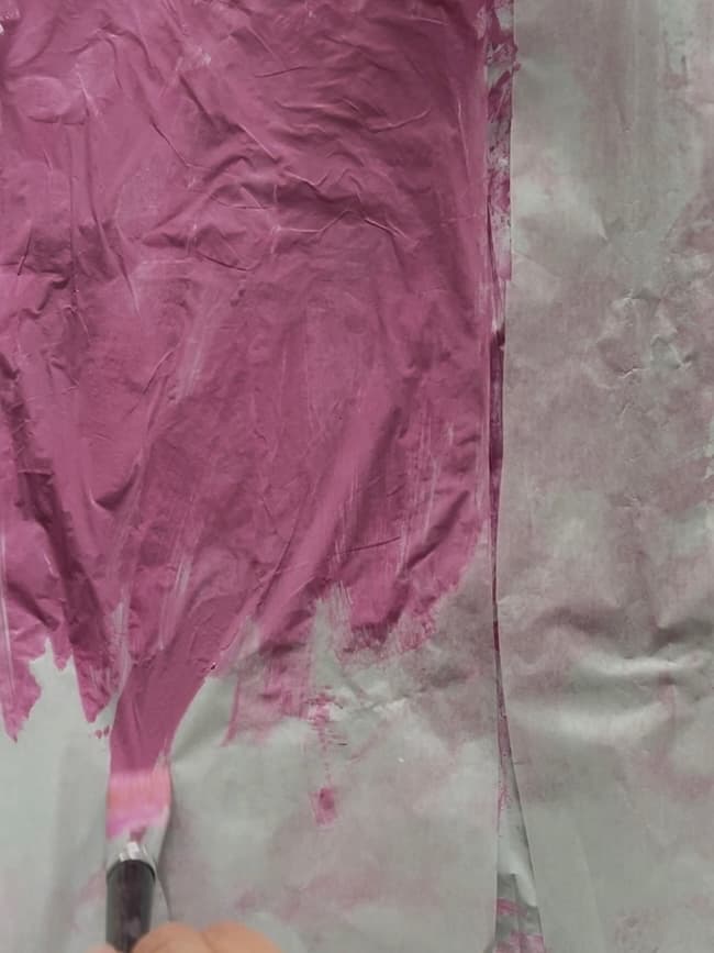 A hand uses a paintbrush to apply pink paint on a surface partially covered with crumpled tissue paper. The paper is textured, resembling the delicate pattern of budding flowering branches as the paint spreads unevenly across it.