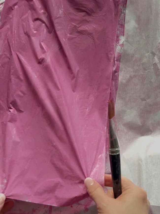 Hands holding a sheet of crumpled pink tissue paper, with one hand using a paintbrush to apply pink paint. The paper has a wrinkled texture reminiscent of flowering branches against the pale background.