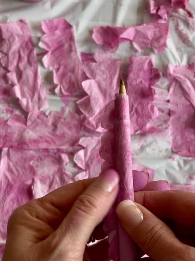 Hands wrapping a pencil with pink floral tissue paper featuring delicate flowering branches. The background displays torn pieces of similar pink tissue paper scattered artfully on a surface.