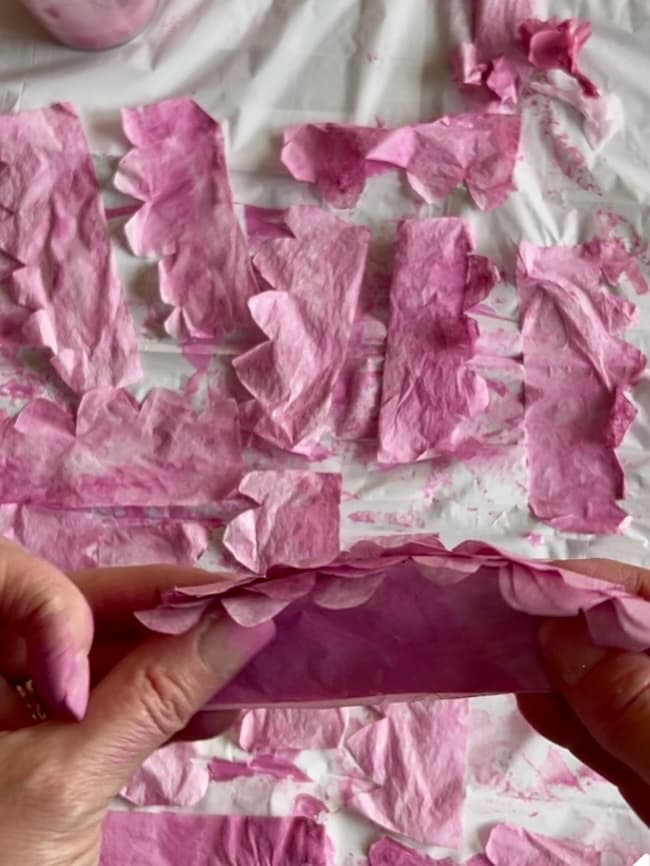 Hands holding a piece of pink tissue paper with scalloped edges evoke the delicate beauty of flowering branches. The background features similar pink strips on a white surface, some slightly crumpled or torn, capturing the natural imperfections found in nature's blossoms.