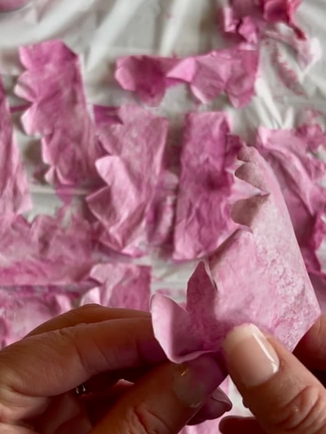 Two hands folding a piece of pink tissue paper, resembling delicate flowering branches, with other ripped pieces spread out on a surface in the background.