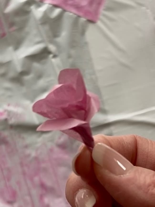 A hand holding a small, delicate pink paper flower crafted from tissue paper against a blurred background. The crumpled texture mimics flowering branches, giving the piece a beautifully handcrafted appearance.