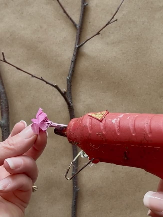 A pair of hands using a red hot glue gun to attach a small pink tissue paper flower to a thin bare branch against a brown background.