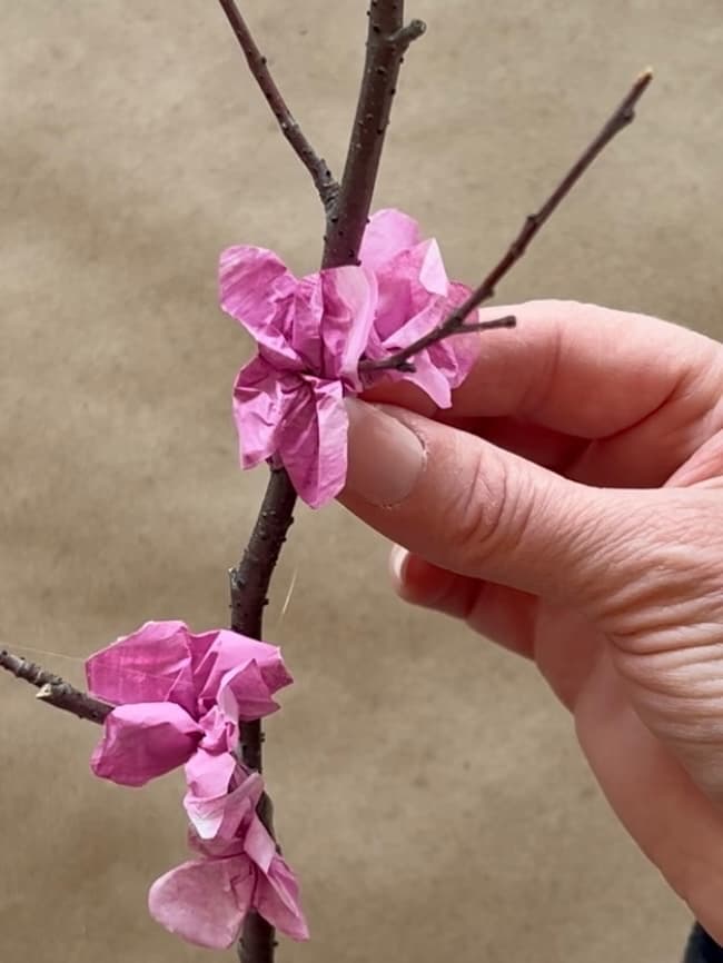 A hand holding a branch decorated with pink, crumpled tissue paper flowers against a beige background.