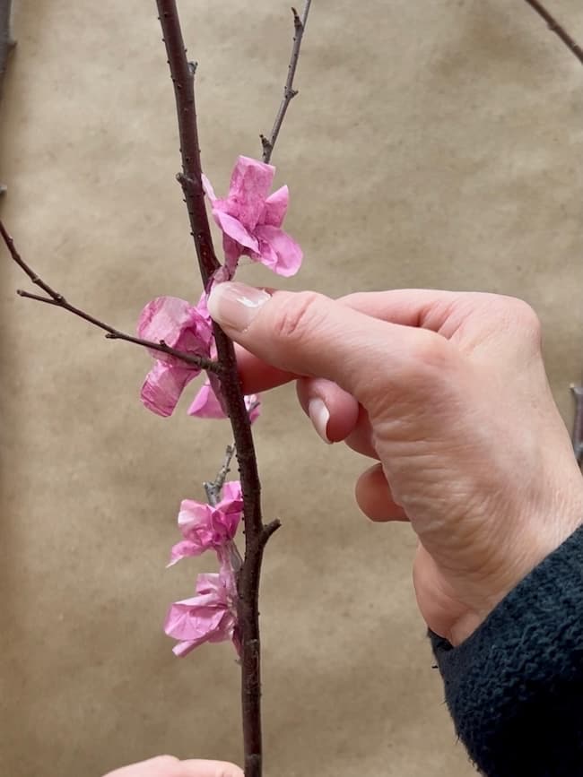 Hands are delicately attaching pink tissue paper flowers to bare branches, transforming them into exquisite flowering branches against a plain background.