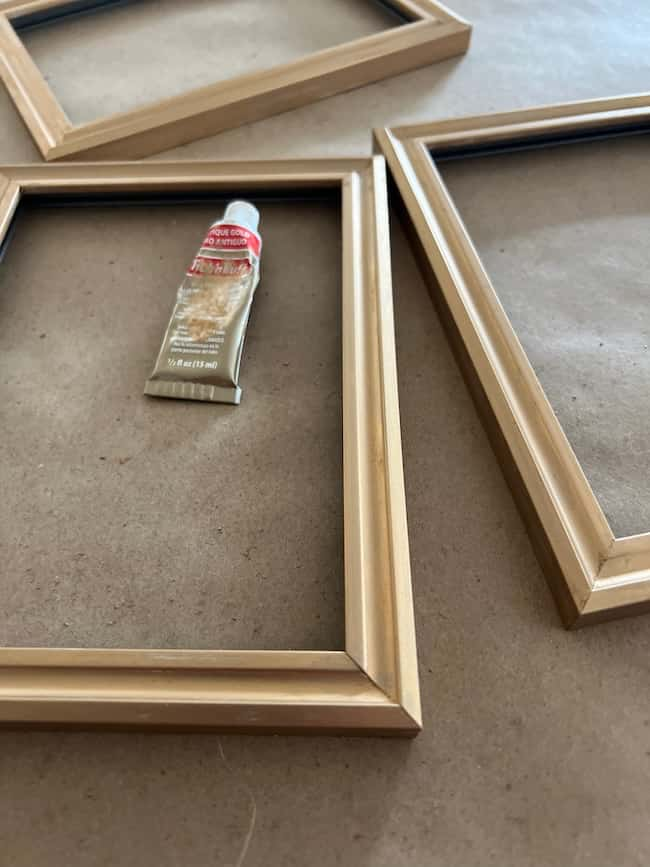 Several wooden picture frames lie on a table, with a small tube of glue placed between them and some free printable art ready to be showcased. The frames are arranged to form a partial square, set against a plain, light brown surface.