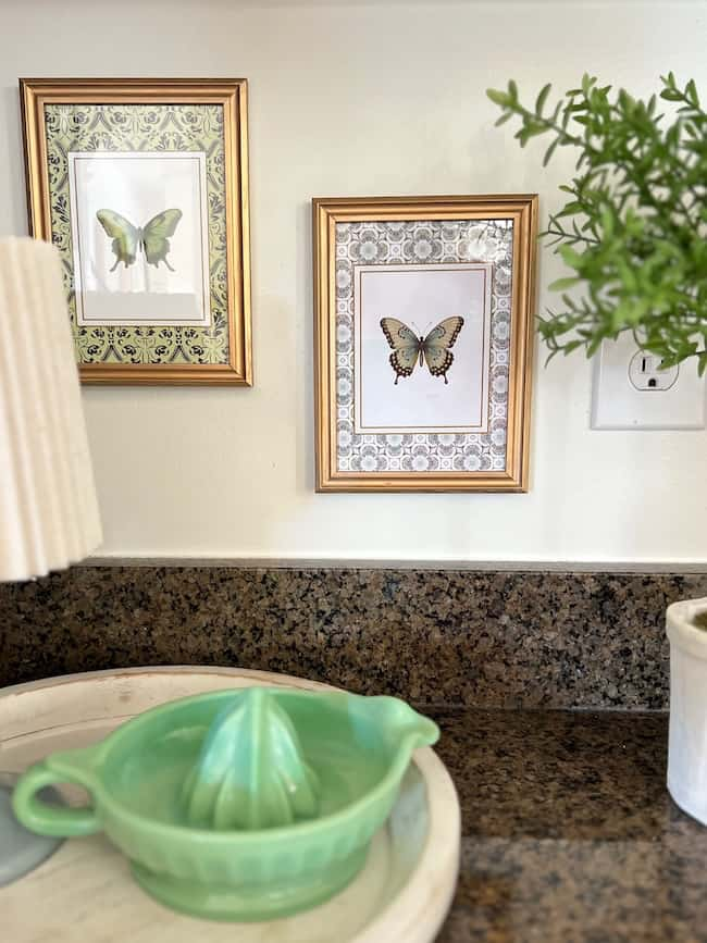 Two framed butterfly prints, reminiscent of free printable art, hang on a light wall with a countertop below. The counter holds a green juicer, a small potted plant, and a lamp with a ribbed shade. An electrical outlet is visible on the wall.