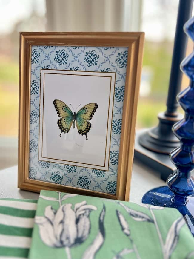 Framed butterfly illustration against a blue floral background, perfectly complementing the free printable art. The frame is gold with a white mat, sitting gracefully on a table beside a blue glass lamp and green and white floral fabric.