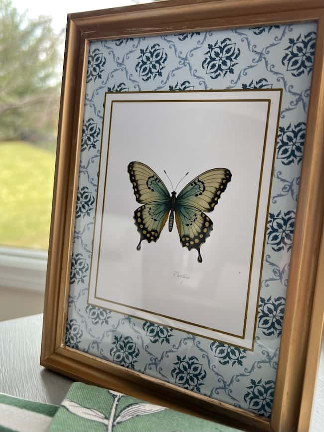 A framed illustration of a butterfly with green and black wings is available as free printable art. It features a decorative blue and white floral patterned border. The gold-colored frame sits on a table by a window, offering a blurred view of the greenery outside.