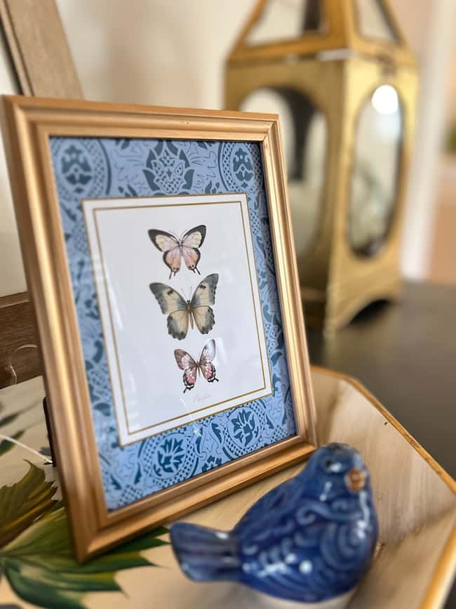 A framed picture of three butterflies with a blue patterned border graces the table, complemented by a ceramic blue bird. In the soft-focus background, a gold lantern adds warmth. This charming setup is inspired by free printable art to elevate any space effortlessly.