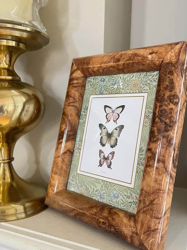 A wooden frame showcasing three butterflies illustrated on free printable art sits next to a brass-colored decorative lamp. The ensemble is beautifully set against a floral-patterned background.