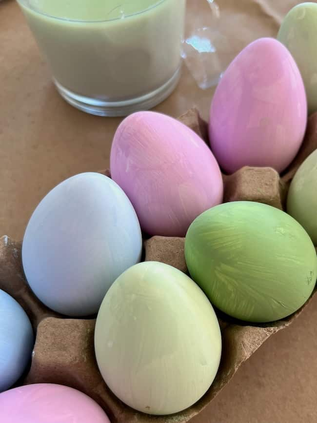 A close-up of hand-painted Easter eggs in a cardboard carton showcases pastel-colored charms. The eggs display soft shades of pink, green, and blue, capturing the essence of Easter decor. A glass with light green paint is partially visible in the background.