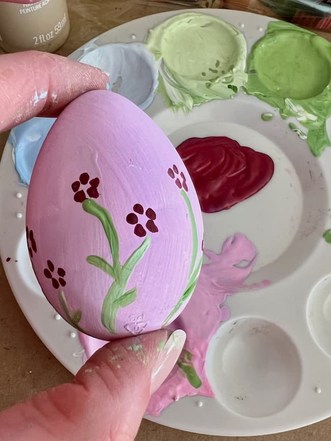 A hand cradles a hand-painted Easter egg adorned with pink and red flower designs nestled among green stems. In the background lies a paint palette showcasing vibrant spring decor hues like blue, green, and red.