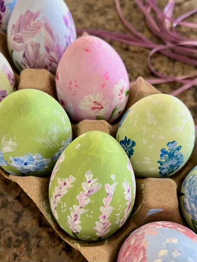 A carton holds intricately painted pastel Easter eggs with floral designs. The hand-painted eggs feature pink, purple, and blue flowers on shades of pink, green, and blue backgrounds. A purple ribbon is partially visible in this spring decor setting.