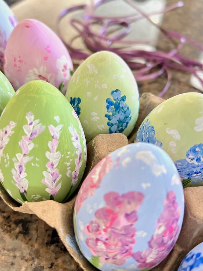 Colorful hand-painted Easter eggs with floral designs are nestled in a cardboard carton. Featuring purple, green, and blue backgrounds adorned with pink and blue flowers, they add a charming touch to any Easter decor. A purple ribbon is softly blurred in the background, enhancing the festive scene.